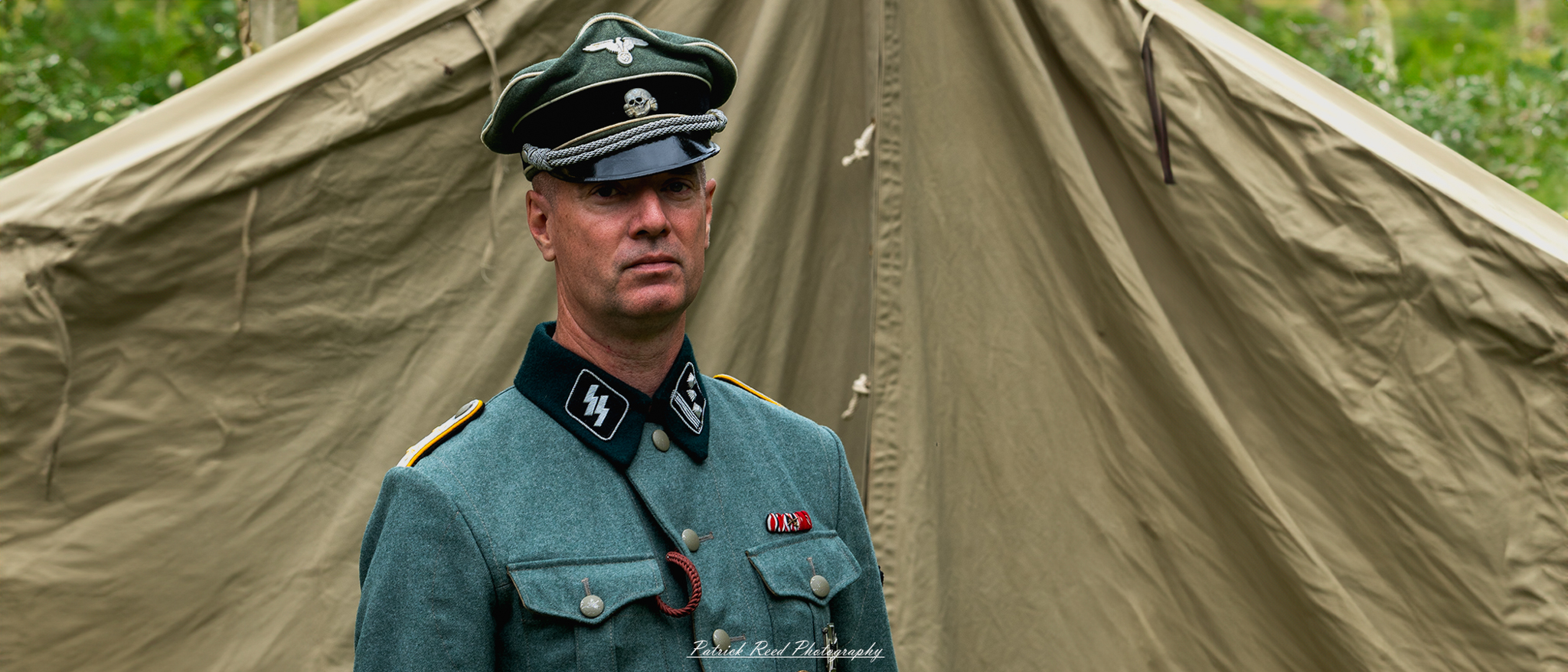 A German officer standing in front of a tent, dressed in a crisp uniform adorned with insignia. His posture is authoritative, and he gazes intently, embodying the leadership and discipline expected in a military setting.